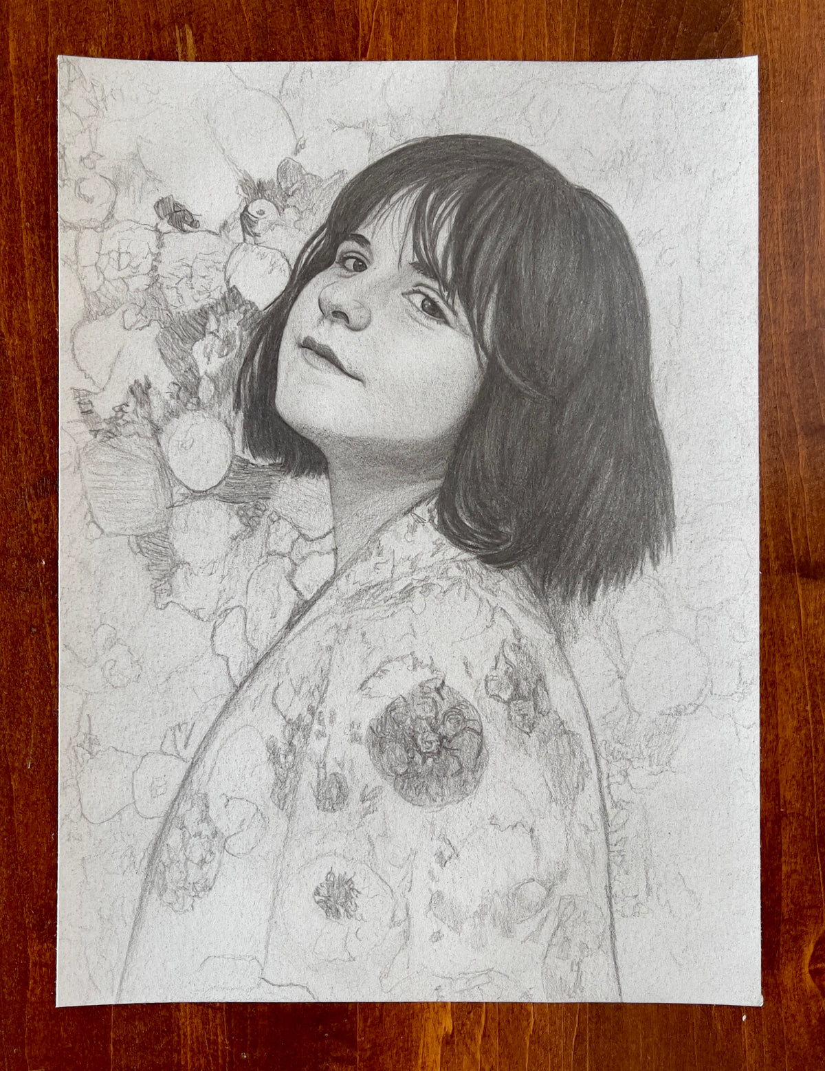 MEFREEK Seth Jennemann Original Pencil Drawing A Fire Escape Made Of Punchlines AKA A Life Jacket Full Of Laughter – Detailed graphite pencil portrait of a young woman with a dark, chin-length bob haircut and wispy bangs, looking over her shoulder toward the viewer with a subtle, thoughtful expression. Her softly shaded face features expressive eyes, delicate freckles, and gently parted lips. She wears a loose, patterned garment decorated with intricate floral motifs, circular ornaments, and abstract illust