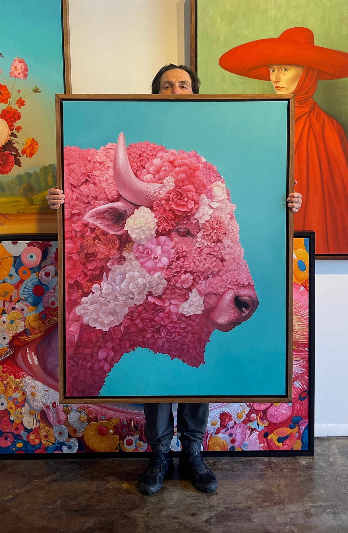 MEFREEK Seth Jennemann Original Oil Painting Armor For A Hopeless Charge artist Seth Jenneman holding a large framed hyperrealistic floral bison portrait composed of layered pink red and white blossoms forming the animal’s face and body against a luminous turquoise background displayed in a studio surrounded by vivid contemporary paintings including floral still lifes and surreal portraiture creating a vibrant gallery atmosphere
