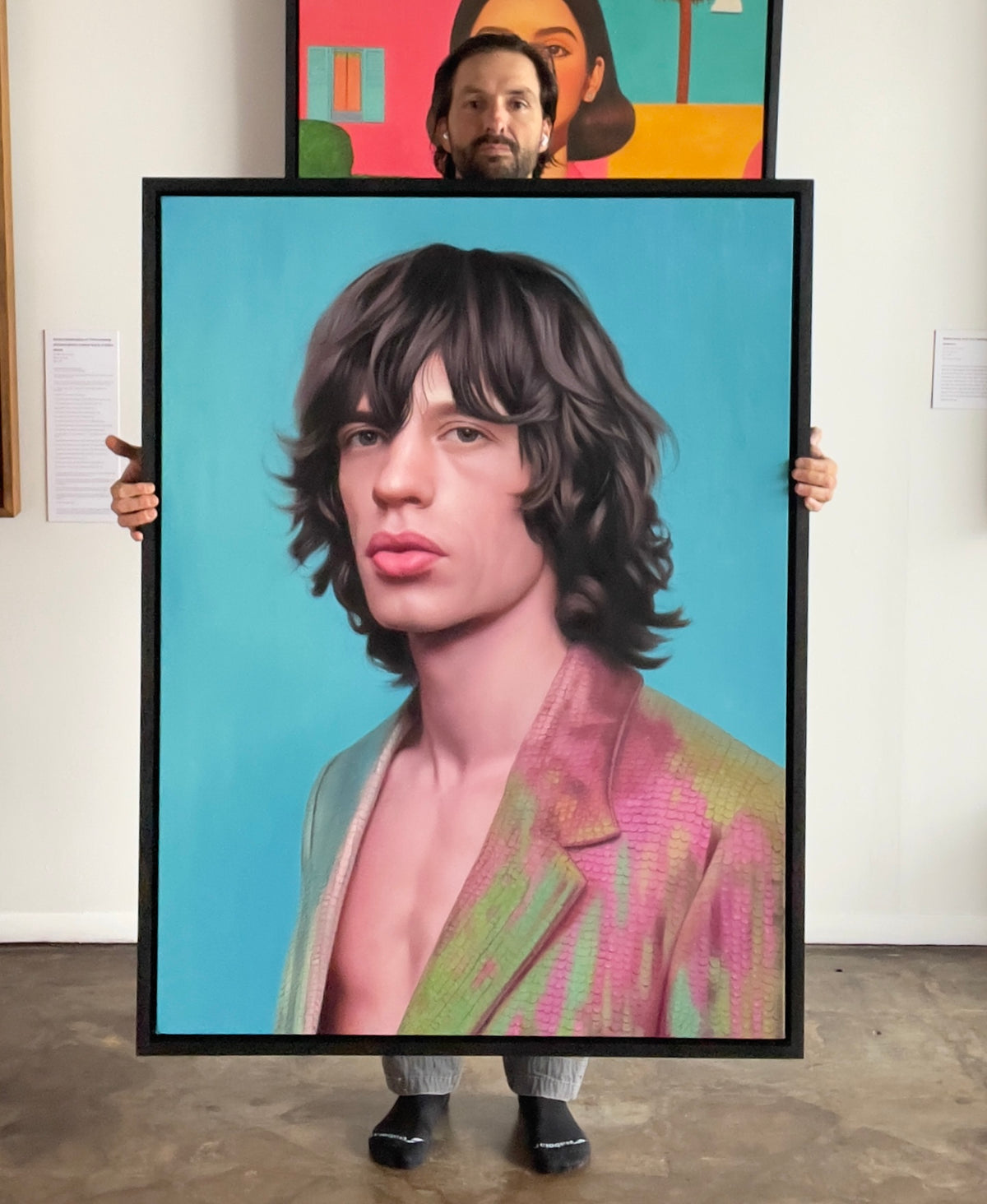 MEFREEK Seth Jennemann Original Oil Painting artist holding a large framed hyperrealistic portrait of Mick Jagger of The Rolling Stones with shaggy dark hair defined cheekbones and signature full lips wearing an iridescent textured jacket in pink green and gold tones set against a smooth turquoise background displayed in a studio with additional colorful paintings visible behind creating contrast between portrait realism and gallery environment