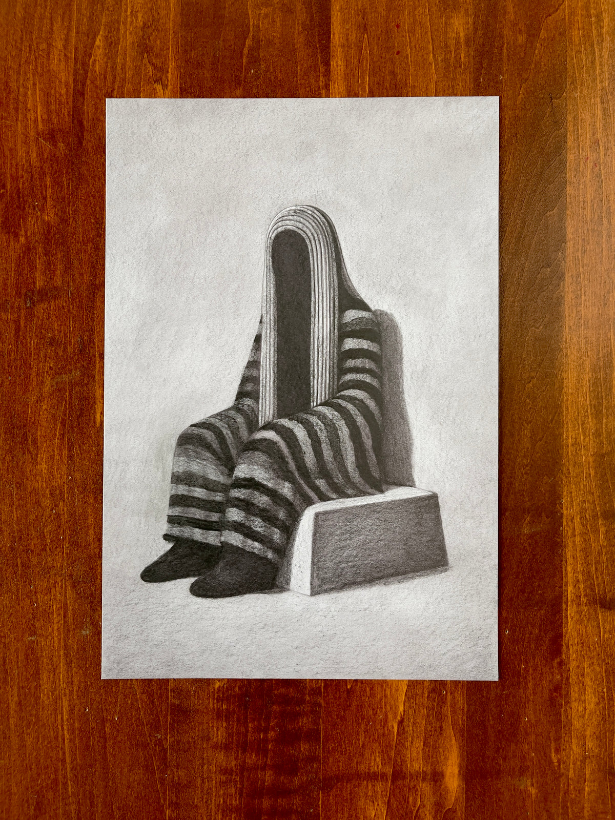 Seth Jennemann MEFREEK — Decoration Draped Over Vulnerability, graphite pencil drawing in hyperreal surrealist style. A solitary faceless figure sits on a block wrapped in striped fabric, rendered with deep tonal contrast and soft gradients that evoke mystery and introspection.