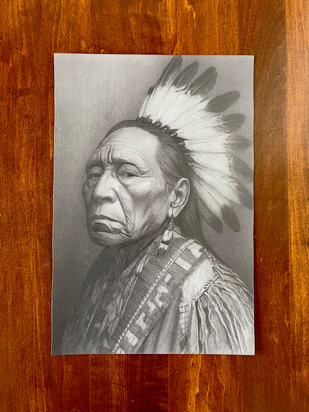 Seth Jennemann MEFREEK Everyone’s To Blame So Nobody’s Responsible – graphite drawing in a hyperreal portrait style depicting an elderly man with a solemn expression, detailed wrinkles, traditional feathered headdress, and intricately patterned garment against a neutral background.
