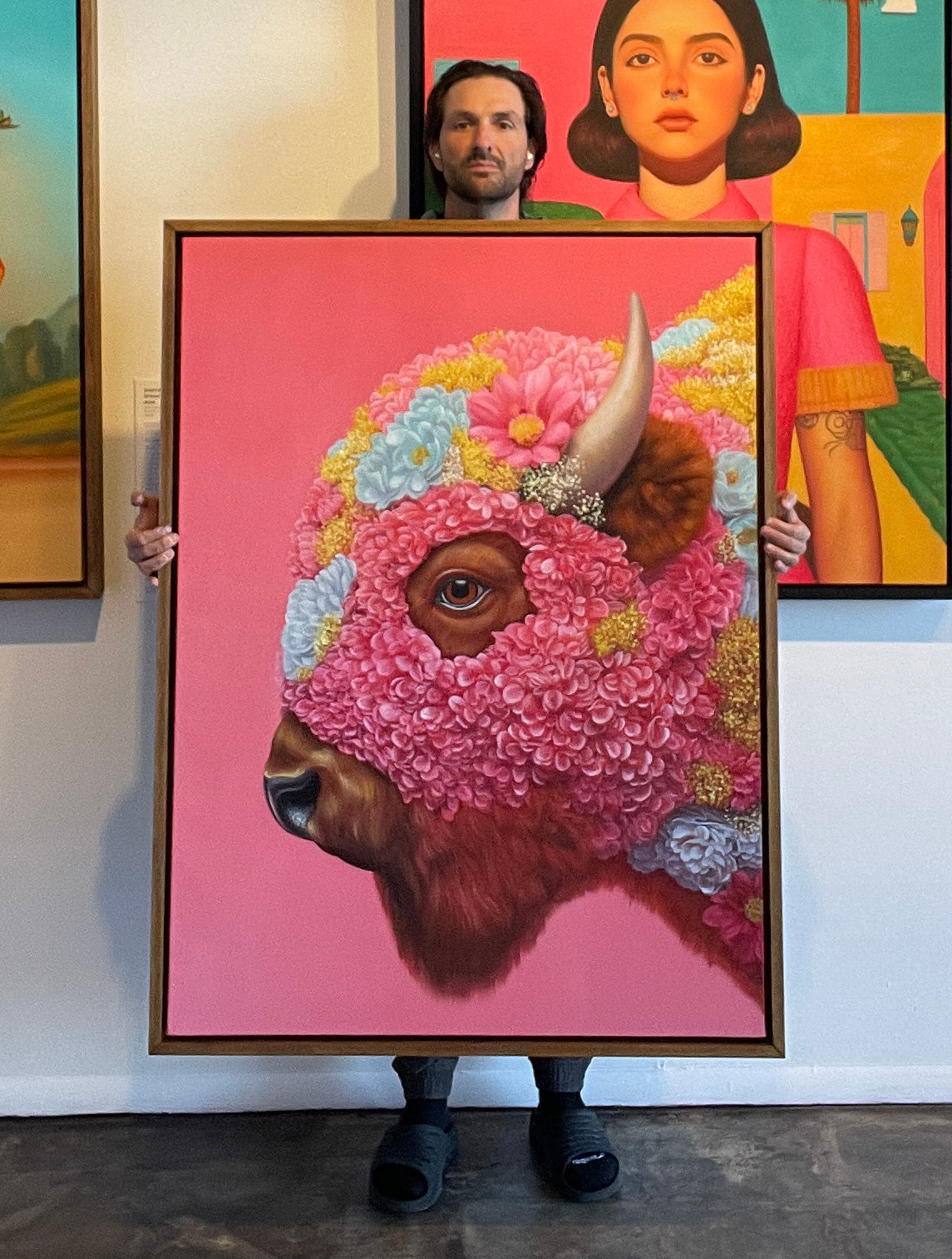 MEFREEK Seth Jennemann I Was Just The Frame artist Seth Jennemann holding a framed hyperreal oil painting of a buffalo wearing a pink floral mask with blue and yellow flowers on a pink background, contemporary surreal realism painting in studio