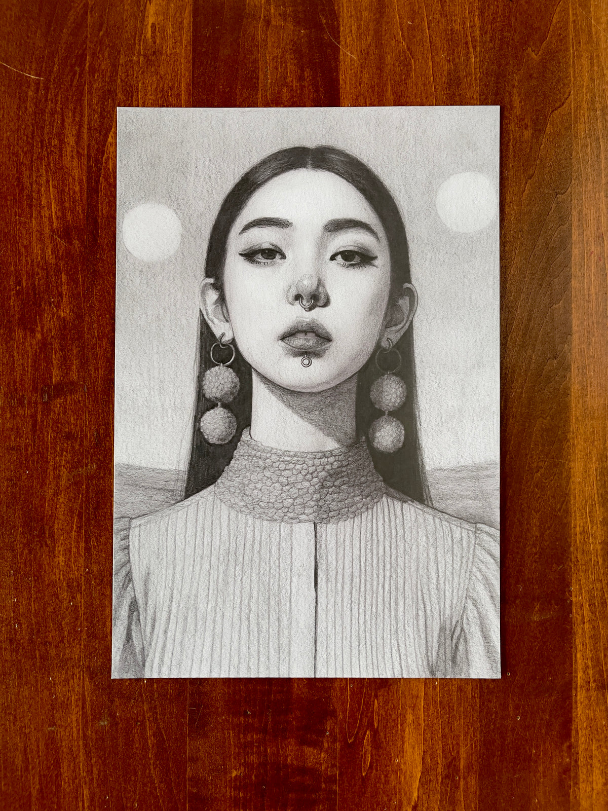 Seth Jennemann MEFREEK I Just Want To Be Me – graphite drawing in a hyperreal contemporary portrait style featuring a serene woman with nose and lip piercings, large textured earrings, and twin moons behind her head, against a soft grayscale landscape background.
