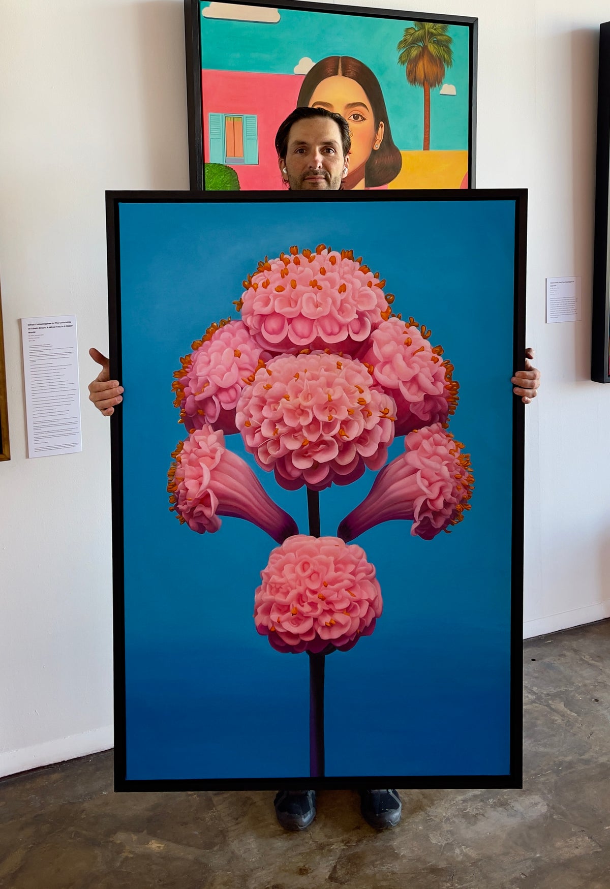 MEFREEK Seth Jennemann It Tastes Like A Compliment If You're Starving artist Seth Jennemann holding large framed hyperreal oil painting of pink sculptural flower forms on a blue gradient background, contemporary surreal botanical painting in studio
