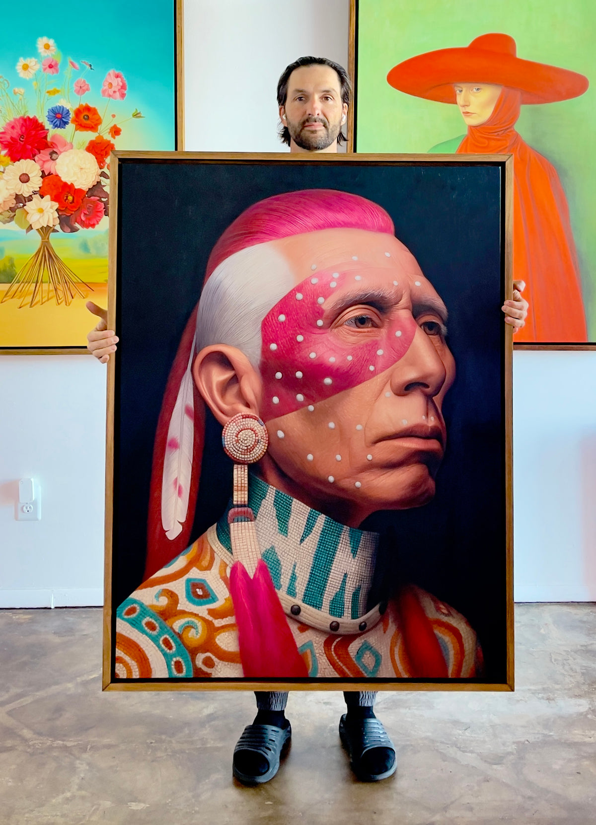 MEFREEK Seth Jennemann Original Oil Painting artist holding a large framed hyperrealistic portrait of a Native American man in profile with vivid pink face paint and white dot markings intricate beadwork earring and patterned garment the painting set against a dark background while the artist stands in a bright studio space with additional colorful paintings behind him including a floral arrangement and a figure in a wide red hat creating contrast between vivid contemporary portraiture and gallery environme