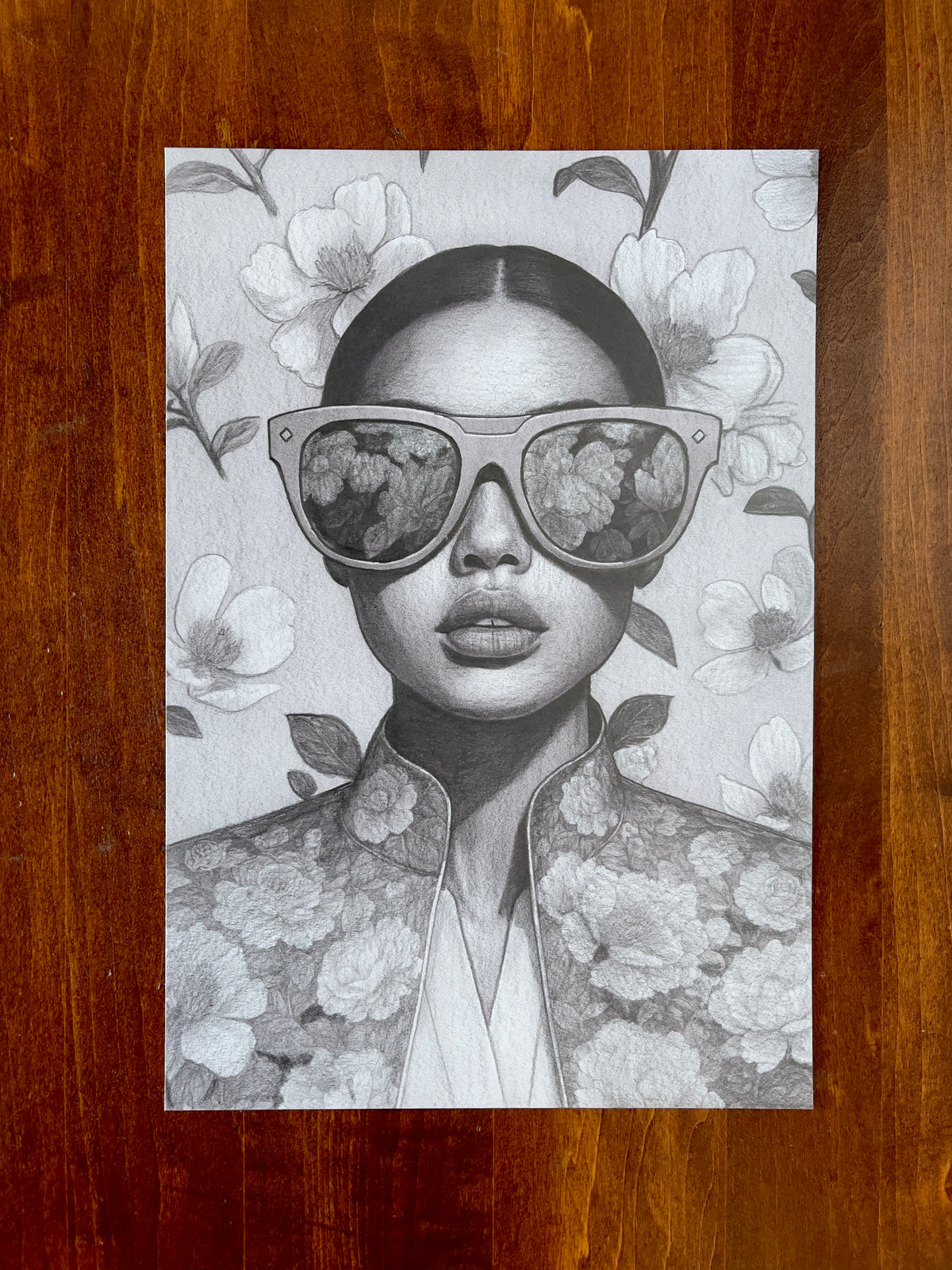Seth Jennemann MEFREEK Now With Less Feeling – graphite drawing in hyperreal pop surrealism style depicting a poised woman in oversized reflective sunglasses and an ornate floral jacket, with blossoms mirrored in her lenses against a softly rendered floral backdrop.
