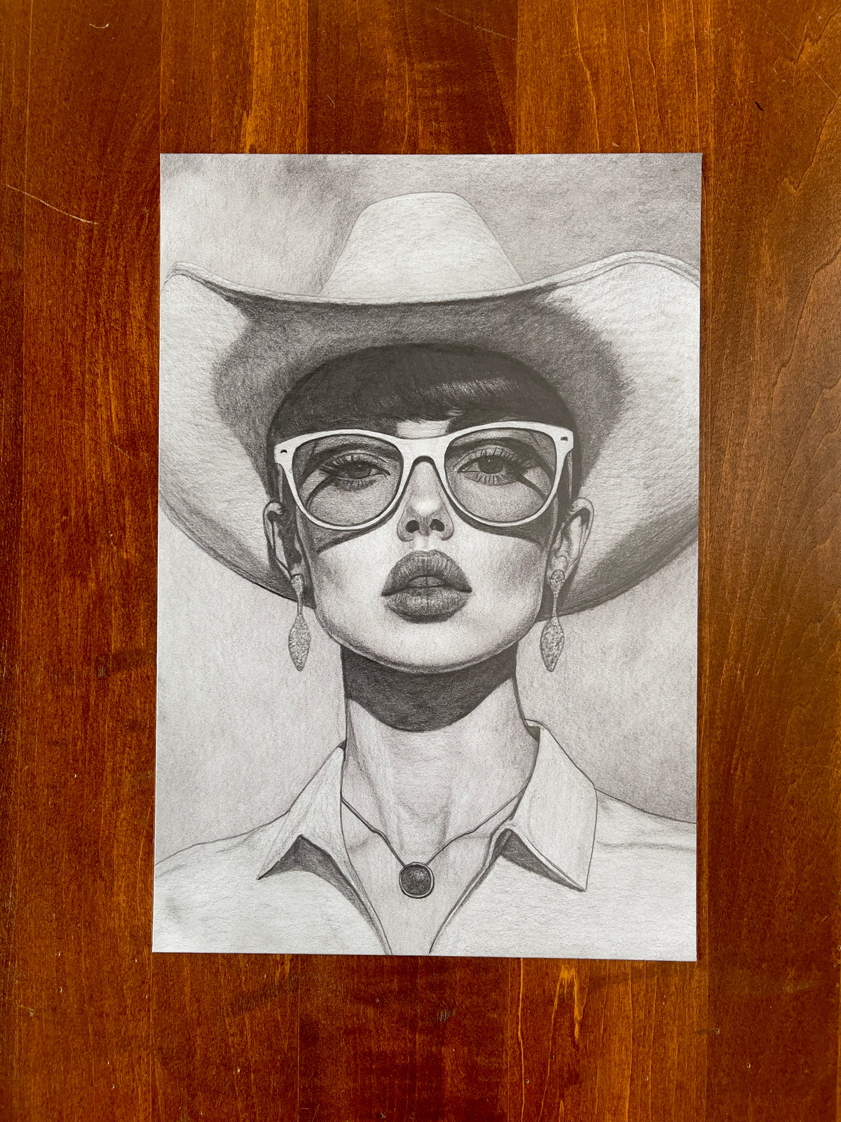 "Rooms Full of Empty People" by Seth Jennemann of MEFREEK, a hyperrealistic graphite pencil drawing of a woman wearing a cowboy hat, large glasses, earrings, and a collared shirt, rendered with precise shading and rich tonal depth on a wooden background.