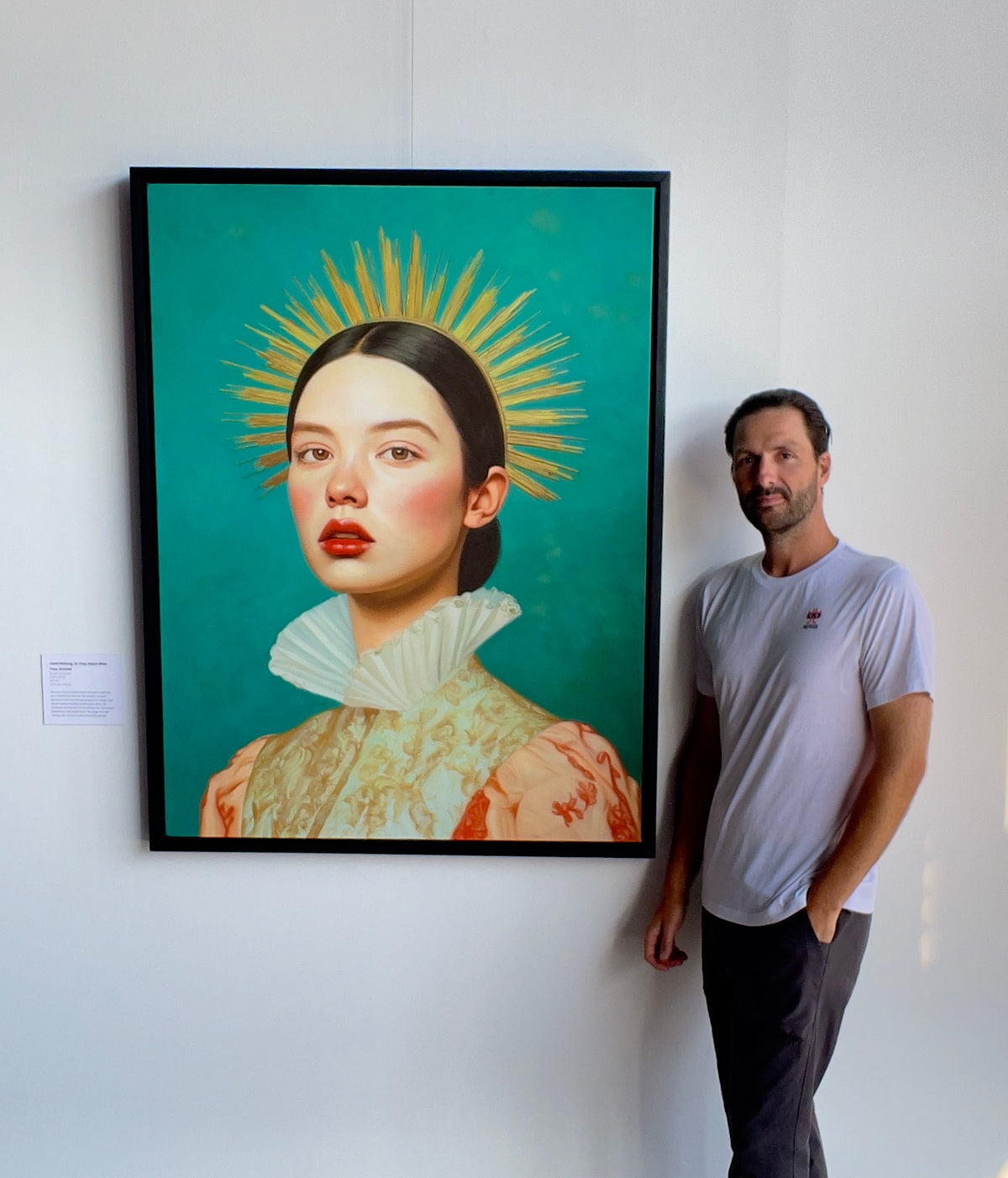 Seth Jennemann MEFREEK I Said Nothing, So They Heard What They Wanted oil painting hyperreal pop surrealism contemporary portrait featuring a solemn woman with dark hair, luminous skin, and a dramatic gold sunburst halo, wearing an ornate patterned gown with a white ruffled collar, shown with Seth Jennemann standing next to the piece in the gallery.
