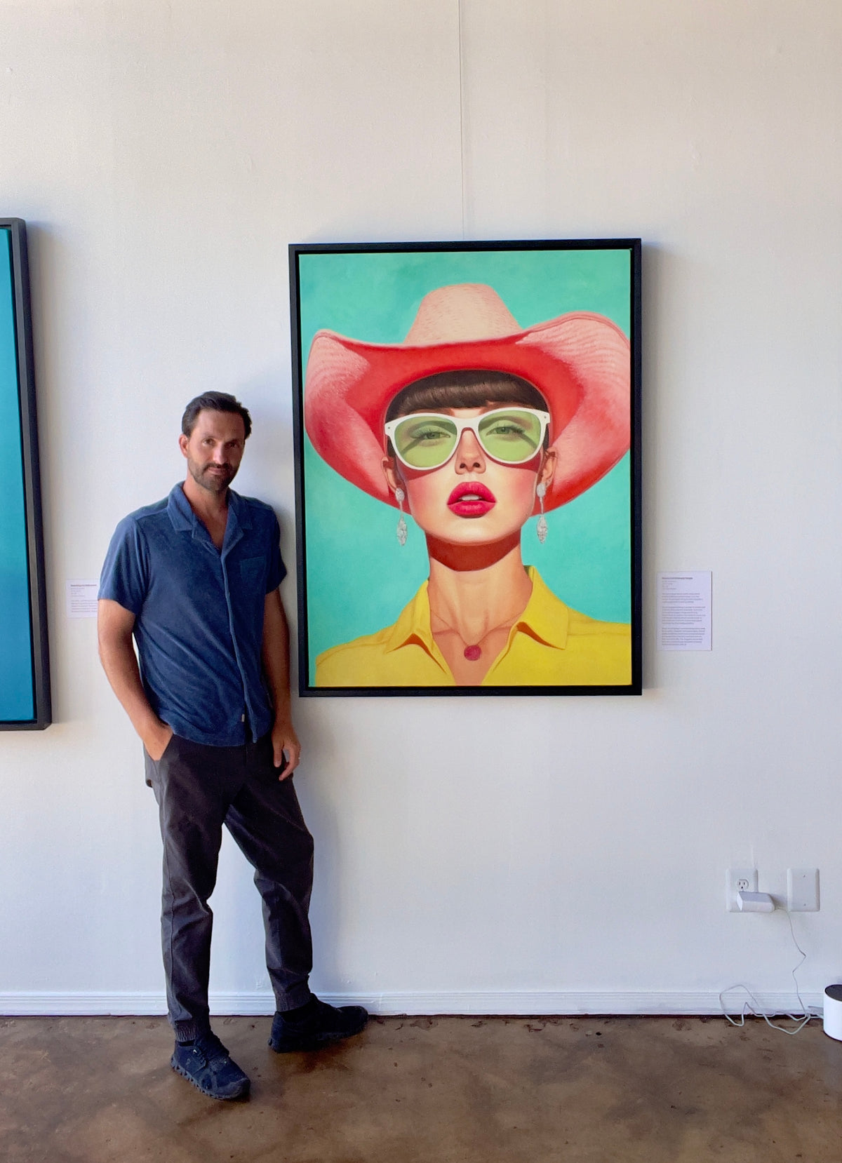 Seth Jennemann MEFREEK Rooms Full Of Empty People oil painting hyperreal pop surrealism contemporary portrait of a glamorous woman in a soft pink cowboy hat, oversized green sunglasses, yellow blouse, and drop earrings, displayed in-studio with artist Seth Jennemann standing beside it against a turquoise background.