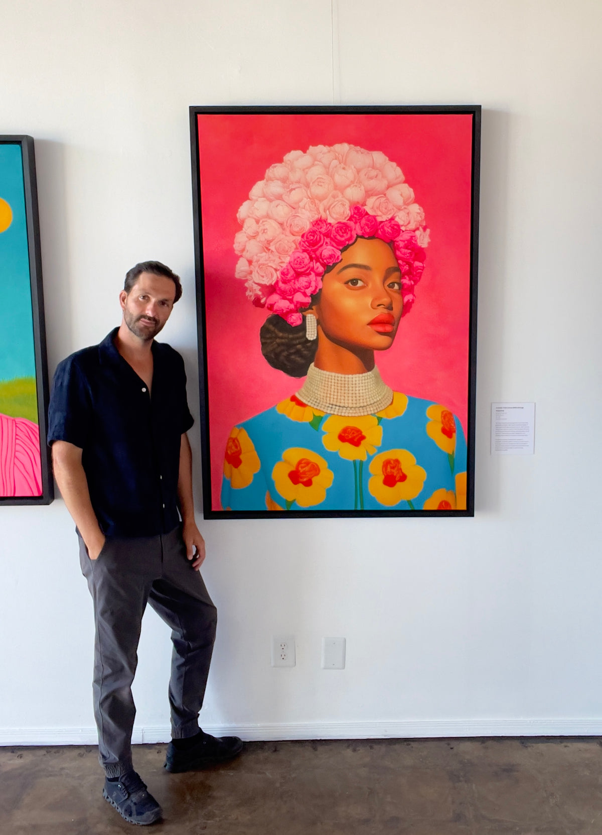 Seth Jennemann MEFREEK A Smile That Came With Strings Attached oil painting hyperreal pop surrealism contemporary portrait featuring a regal woman with a towering crown of pink roses, pearl earrings, and a pearl collar, dressed in a blue garment adorned with bold yellow flowers, shown with Seth Jennemann standing next to it in the gallery.