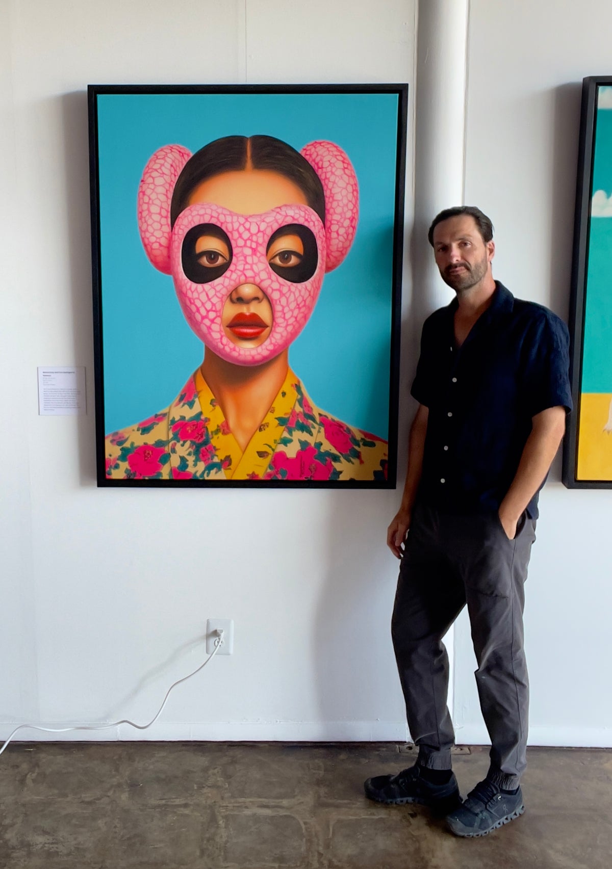 Seth Jennemann MEFREEK Melancholy And The Fleshlight Of Sadness oil painting hyperreal pop surrealism contemporary portrait featuring a woman wearing a textured pink mask with large ear-like protrusions and black eye cutouts, dressed in a floral yellow-collared garment against a vivid turquoise background, shown with Seth Jennemann standing beside the artwork in the gallery.