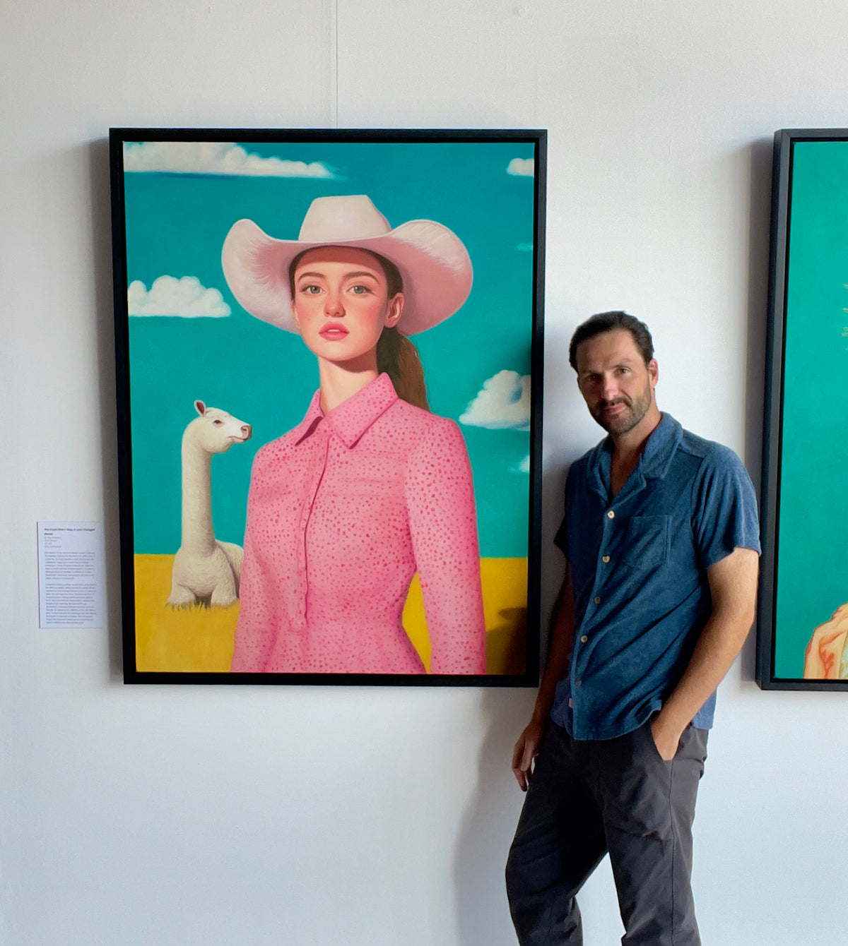 Seth Jennemann MEFREEK The Clock Didn't Stop It Just Changed Hands oil painting hyperreal pop surrealism contemporary portrait of a woman in a pink patterned dress and white cowboy hat standing in a golden field with a white llama, under a bright blue sky with fluffy clouds, shown with Seth Jennemann beside it in the gallery.
