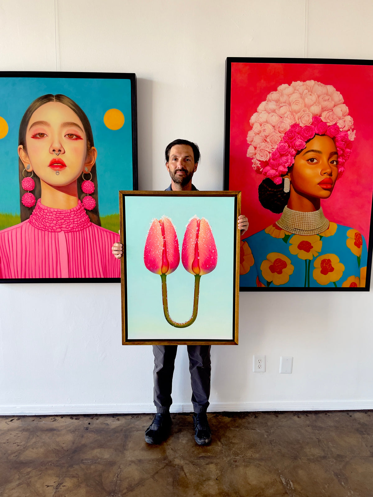 Seth Jennemann MEFREEK A Tether Doesn't Mean You're Connected oil painting hyperreal contemporary floral surrealism shown held by Seth Jennemann in his studio, standing between two large vivid portraits one with pink pom earrings and another with a floral headpiece on bright backgrounds.