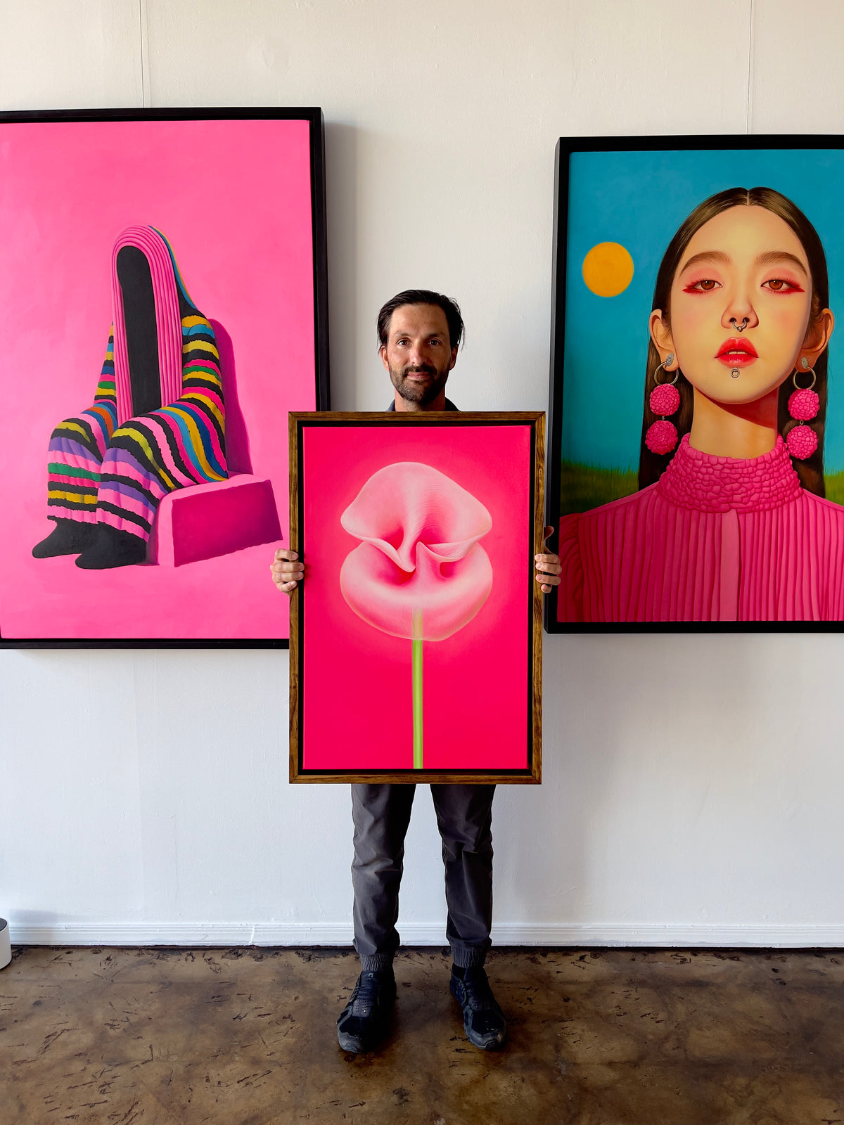 Seth Jennemann MEFREEK An Inheritance That Feels Like A Debt oil painting contemporary surrealism showing Seth Jennemann holding a framed pink abstract floral painting, standing between two large canvases one of a faceless striped figure on the left and one of a woman with pink pom earrings on the right, against a studio wall with concrete floor.