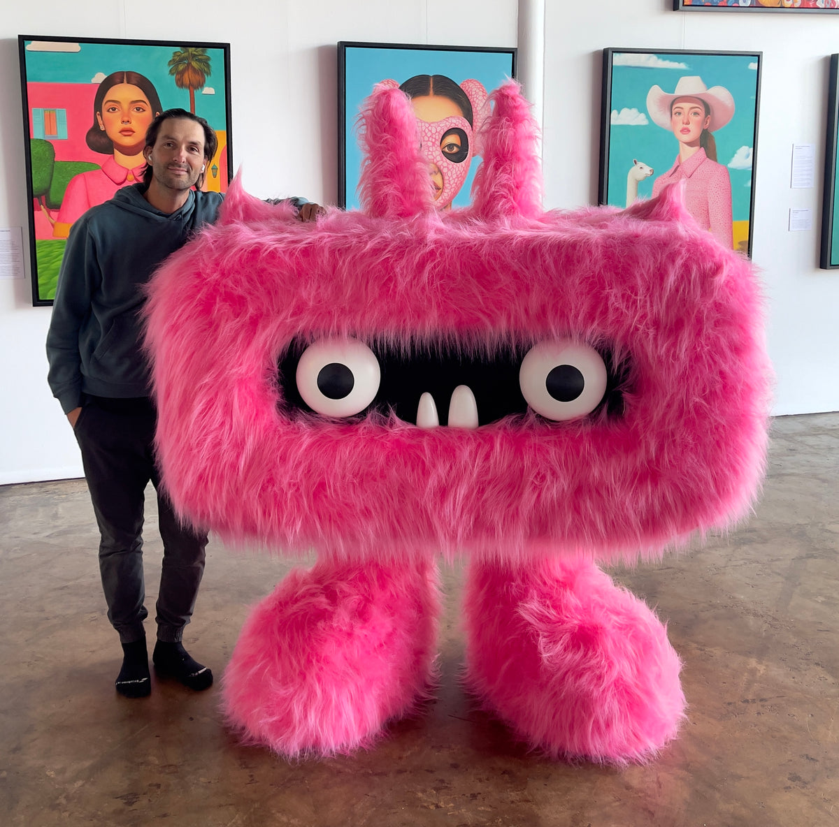 MEFREEK Seth Jennemann with Underbite standing beside a giant pink furry Underbite sculpture at the MEFREEK studio in Tulsa, surrounded by colorful original paintings from the MEFREEK creative universe