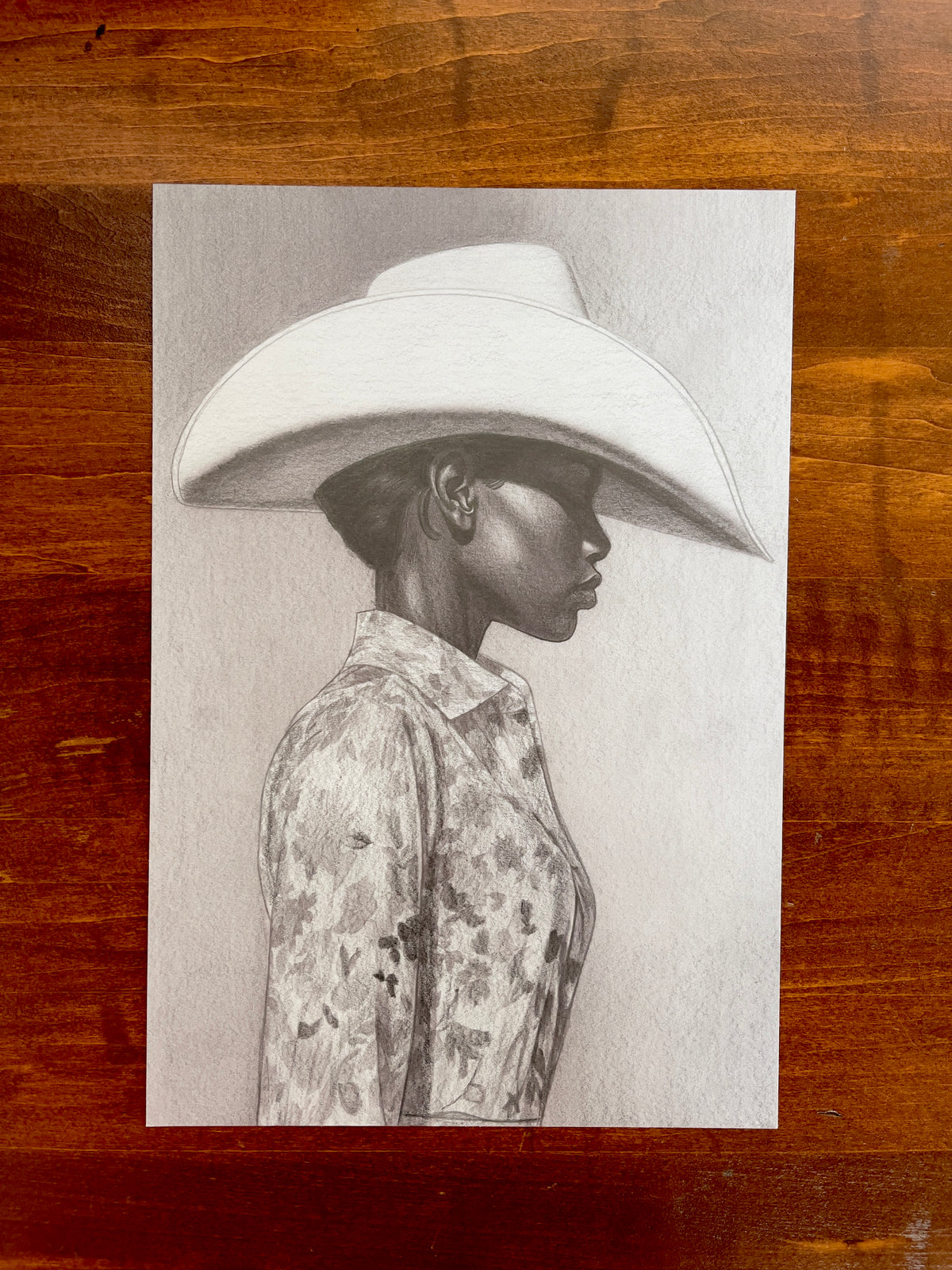 Seth Jennemann MEFREEK The Milk Tastes Funny When You’re On The Carton – graphite drawing in a hyperreal contemporary portrait style showing a side profile of a woman wearing a wide-brimmed cowboy hat and a floral-patterned shirt, rendered with smooth tonal shading and precise detail.
