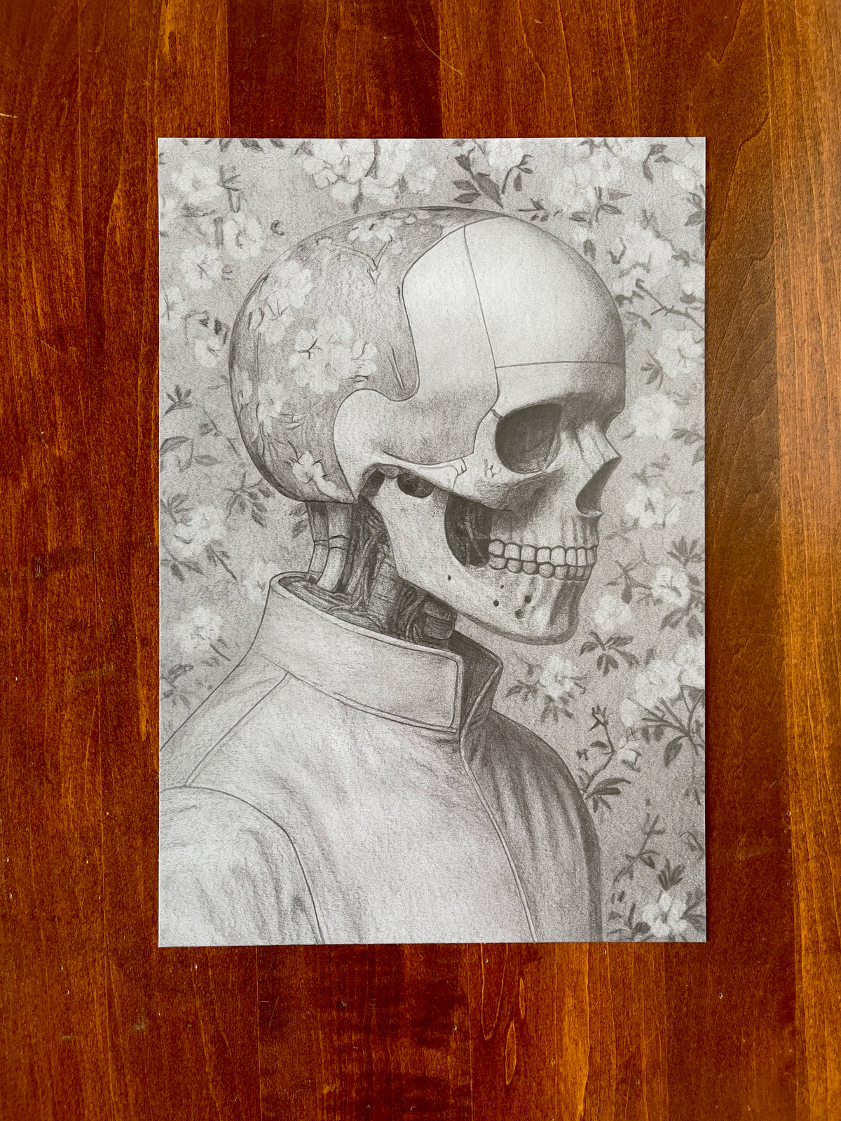 Seth Jennemann MEFREEK The Museum Of The Human Interval – graphite drawing in a hyperreal pop surrealism style depicting a human skull wearing a sleek collared jacket against a delicate floral patterned backdrop, merging mortality with fashion in striking monochrome detail.
