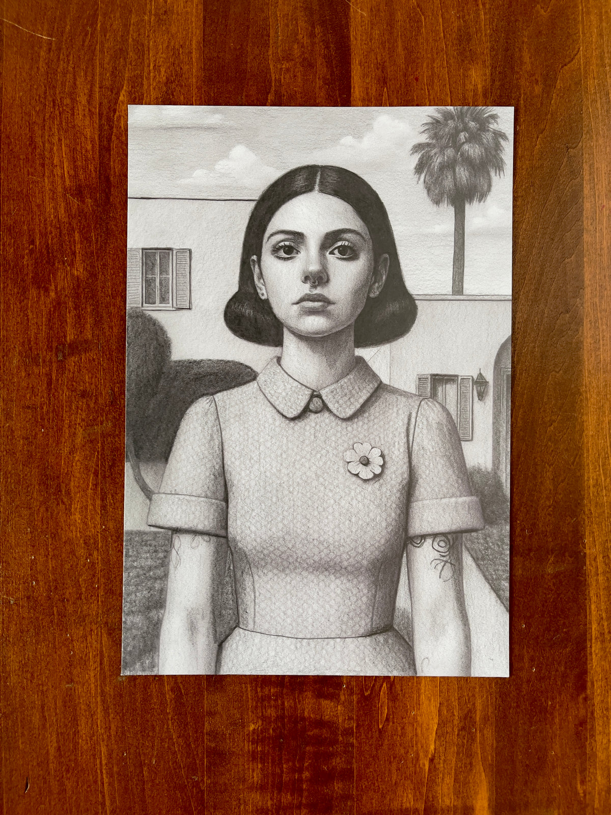 Seth Jennemann MEFREEK — This Only Ends When You Do, graphite pencil drawing in hyperreal contemporary portrait style. A young woman with dark bobbed hair, a collared dress, and a flower pin stands expressionless in a suburban setting with a palm tree and clouds in the background.