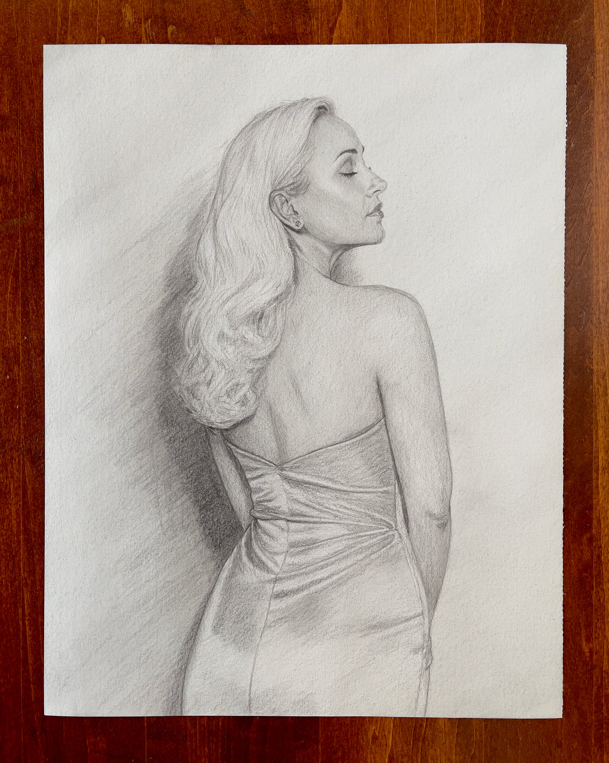 MEFREEK Seth Jennemann Original Pencil Drawing Two For The Price Of Two – Realistic graphite pencil portrait of a woman viewed from behind, turning her head to the right in an elegant side profile with her eyes closed. Her long, voluminous wavy hair cascades over one shoulder, revealing a small stud earring, graceful neck, and bare upper back. She wears a strapless fitted gown with intricately gathered and ruched fabric across the back and waist. Delicate graphite shading captures the soft texture of her ha