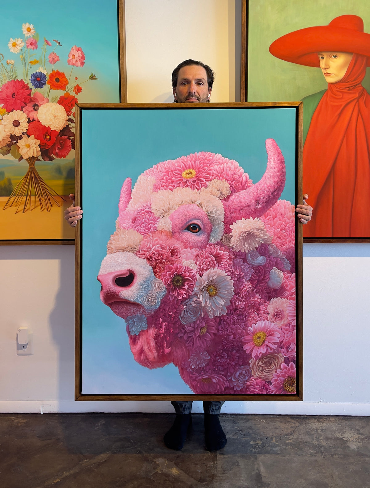 MEFREEK Seth Jennemann Original Oil Painting We Decorated The Cage Ourselves artist jenneman holding a large framed hyperrealistic floral bison portrait composed of layered pink and white blossoms with detailed horns expressive eye and soft organic textures against a luminous turquoise background displayed in a contemporary studio surrounded by vivid floral and surreal portrait paintings creating a colorful immersive gallery atmosphere