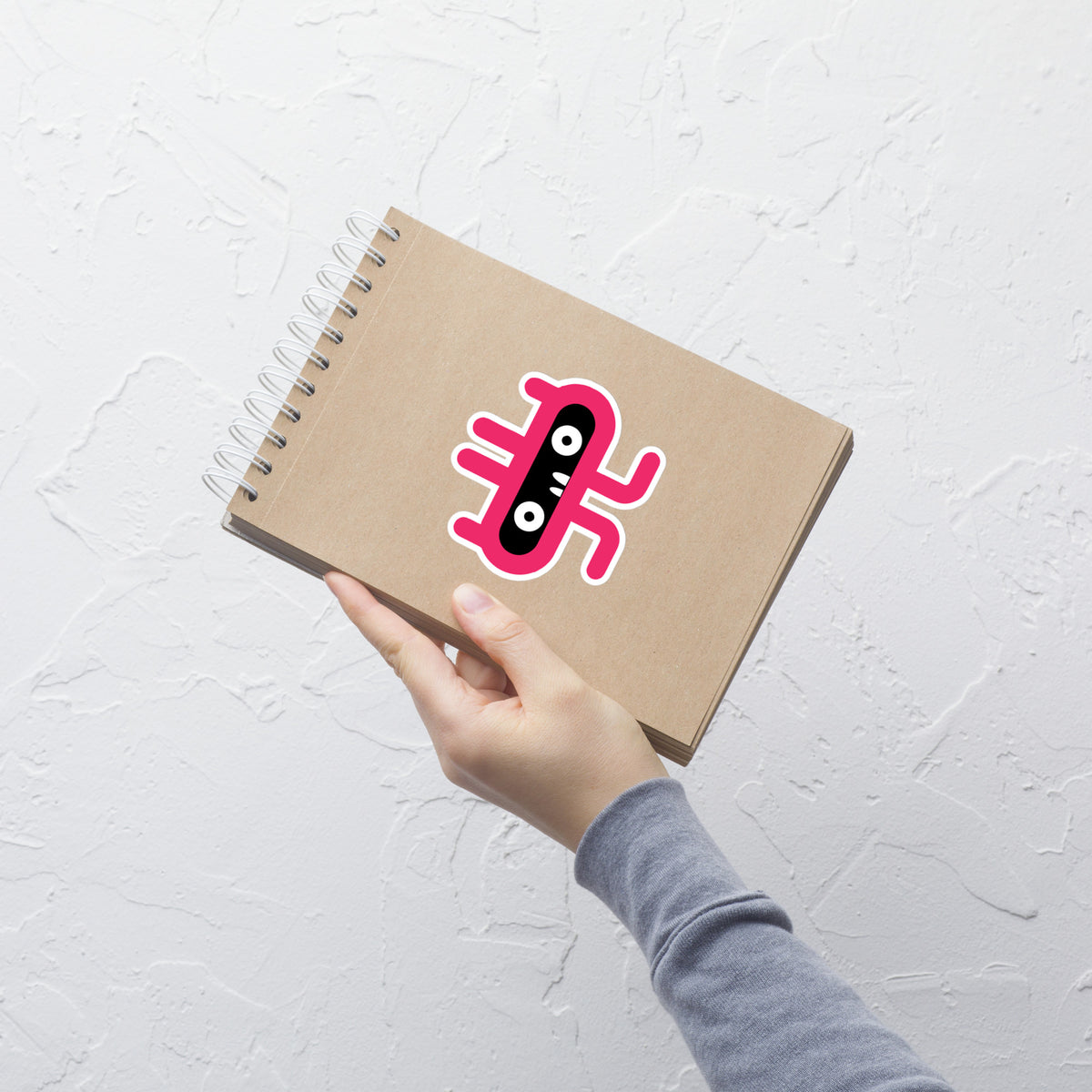 Hand holding a brown notebook with a colorful sticker on a light gray textured background