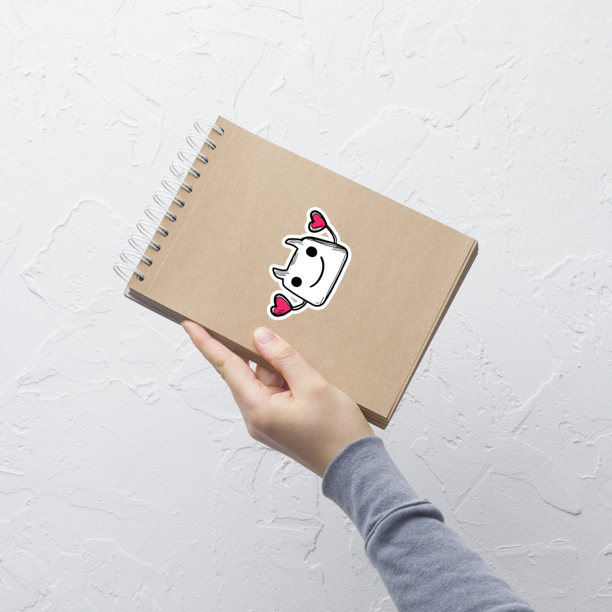 Hand holding a brown notebook with a cartoon character sticker on a light gray textured background