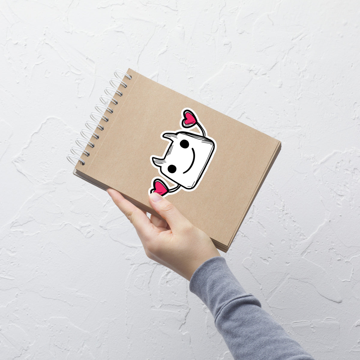 Hand holding a brown notebook with a cartoon ghost sticker on a white textured background