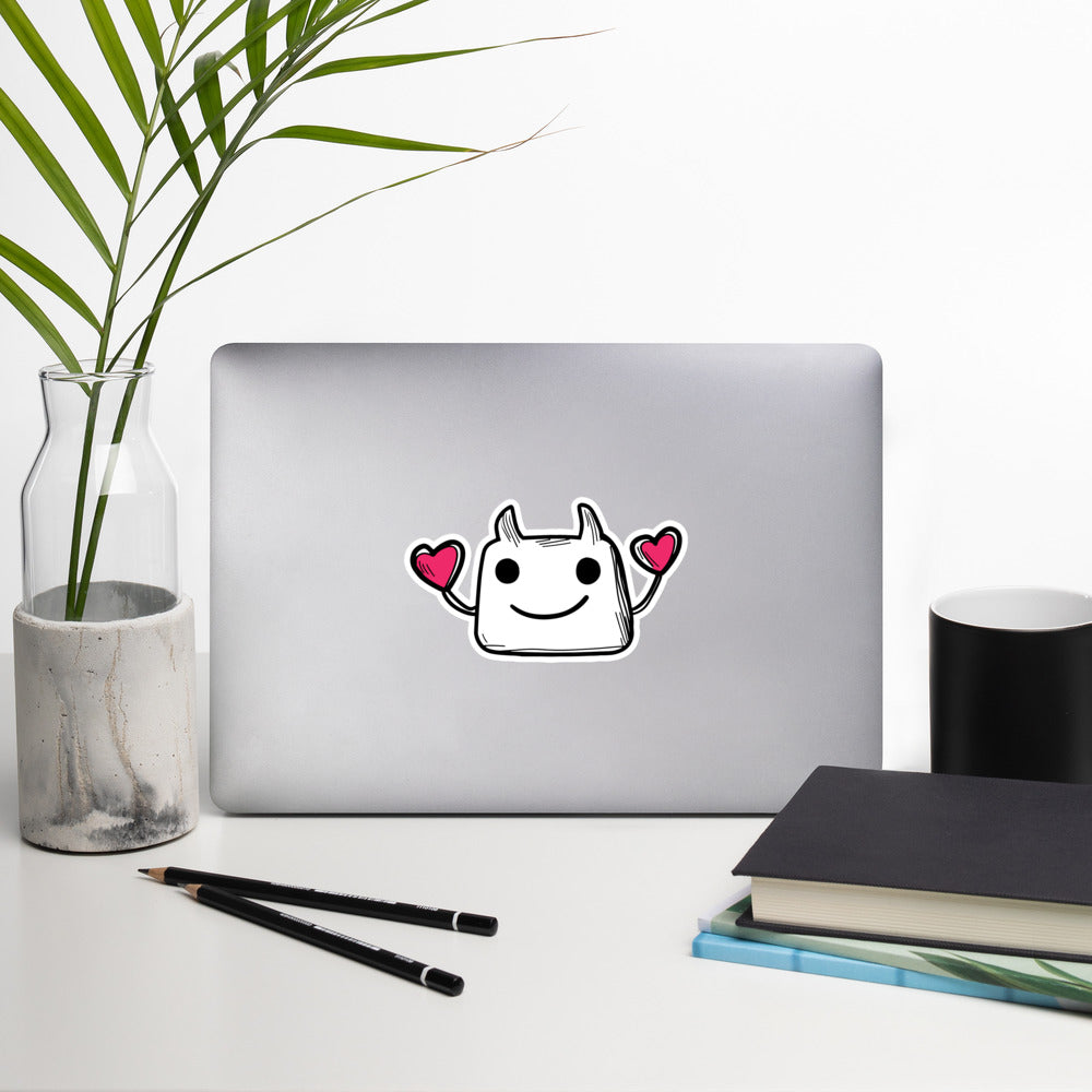 Laptop with colorful sticker on a white surface with a plant, notebook, and pencils.